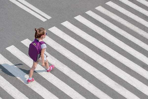 Young girl wearing a purple backpack walking across a wide crosswalk on a sunny day.