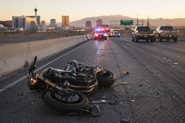 A black motorcycle lying on its side on a Las Vegas highway at dusk with debris scattered on the pavement and emergency vehicle lights flashing in the background, illustrating why Las Vegas roads are considered dangerous for motorcycle riders.